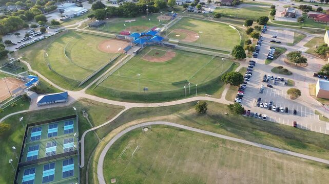 Full tennis pickleball courts with players, baseball softball fields cloverleaf with central blue-roofed pavilion, bleachers, paved paths. Adjacent to parking, trees, light commercial buildings