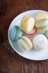 Tender multicolored macaroons with different tastes on white plate, wooden old table background, top view closeup shot