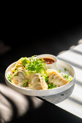 Steamed dumplings served on a white plate with soy dipping sauce, fresh greens, herbs, and chili flakes. Sunlight enhances the appetizing, rustic food presentation.
