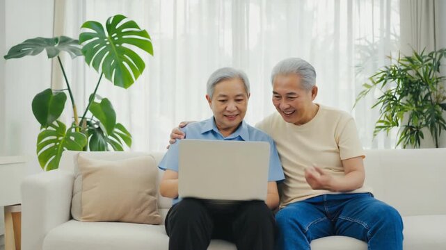 Elderly couple enjoys sharing moments while browsing on a laptop at home in bright, cozy living room