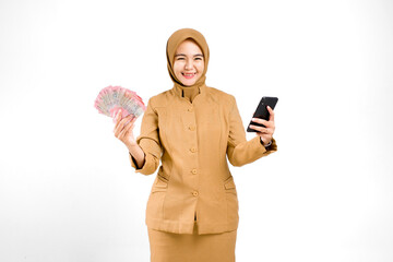 Cheerful young beautiful civil servant in brown uniform showing Indonesian rupiah and smartphone,...
