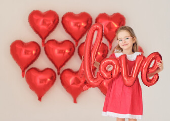 Smiling girl in red dress holds shiny balloon word &ldquo;love&rdquo;.
Heart balloons create festive Valentine background.
Playful studio portrait captures childhood joy and romance theme.
