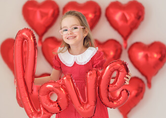 Smiling girl in red polka-dot dress holds balloon word &ldquo;love&rdquo;.
Heart balloons create bright Valentine background.
Joyful studio portrait captures childhood happiness and charm.

