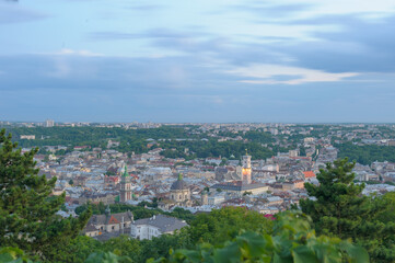 Obraz premium View of Lviv city center from a hilltop at dusk, with blurred sky and city lights glowing, trees in the foreground.