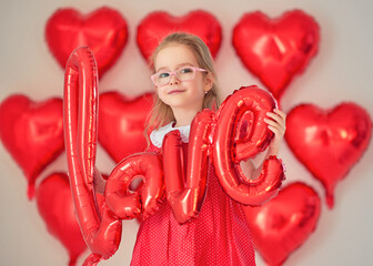 Smiling girl in red dress holds shiny balloon word &ldquo;love&rdquo;.
Heart balloons create bright Valentine studio background.
Playful portrait captures childhood joy and romantic mood.
