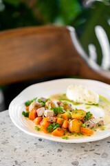 A plate of delicious, hearty stew with chunks of meat, roasted potatoes, and carrots, garnished with fresh herbs and a creamy sauce on a modern restaurant table.