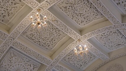 Carved decorative ceiling with two chandeliers. The ceiling features ornate, carved plasterwork, also known as a coffered ceiling or false ceiling, with intricate floral and geometric patterns. - Powered by Adobe