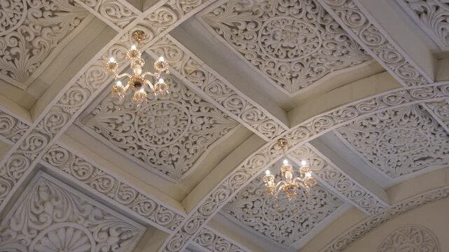 Carved decorative ceiling with two chandeliers. The ceiling features ornate, carved plasterwork, also known as a coffered ceiling or false ceiling, with intricate floral and geometric patterns.
