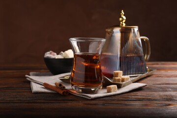 Tasty Turkish tea in glass cup, brown sugar, cinnamon, sweets and teapot on wooden table against color background, closeup
