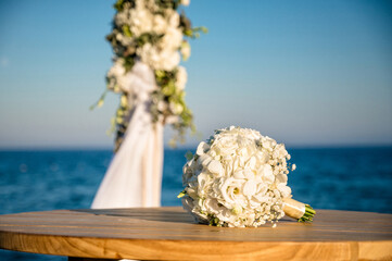 Elegant wedding bouquet of white roses and hydrangeas resting on wooden table by sea under blue sky. Romantic beach ceremony detail with soft natural light and floral beauty.