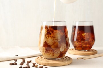 Pouring milk into glass of iced coffee at white table, closeup