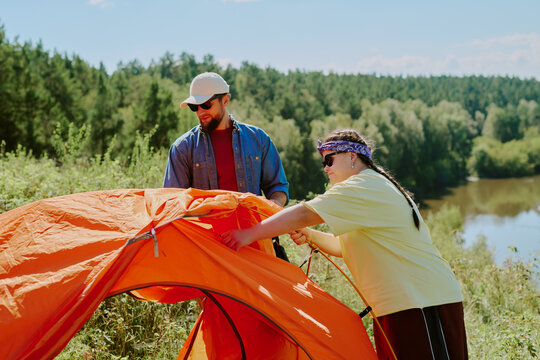 Caucasian young adult man and Caucasian young girl with Down syndrome setting up orange tent outdoors near forest and river, both focused on assembling camping equipment during summer adventure - Powered by Adobe