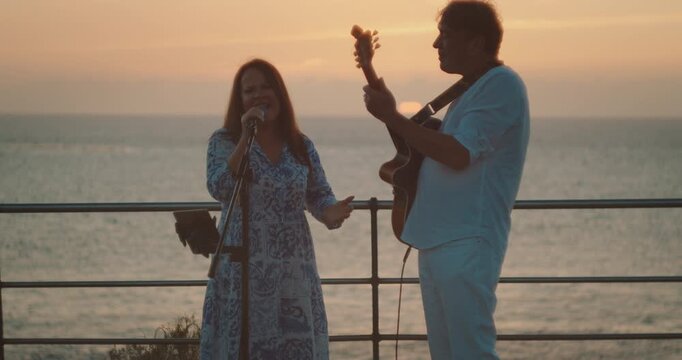 Man playing guitar while woman sings at oceanfront during colorful sunset
