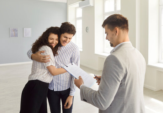 Young happy married couple hugging in new house standing with smiling man realtor and ready to become new homeowners. Professional real estate agent holding contract make a deal with customers