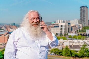 Elderly man with long white beard talking on mobile phone against city skyline with modern buildings on sunny day