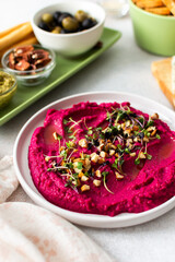 Beetroot hummus is placed in the center of a charcuterie board, surrounded by snacks like olives, nuts, cheese, and crispy breadsticks. A light, inviting setup showcases vibrant colors