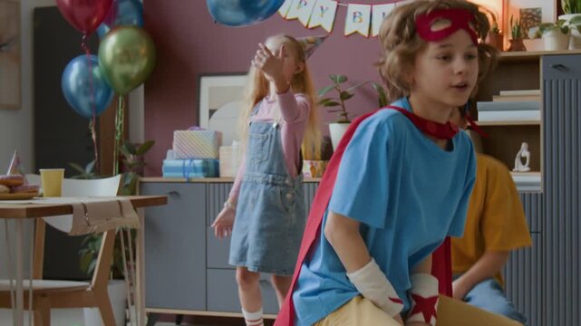Tilt-down shot of two young boys in festive costumes riding bouncing balls while girl playing with balloon during birthday party