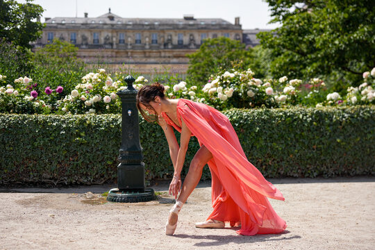 Ballet dancer in coral dress putting on pointe shoes in Paris garden