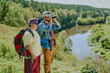 Caucasian young girl with Down syndrome and young man standing on grassy hill overlooking river, man using binoculars while girl observing landscape, both carrying backpacks, surrounded by forest