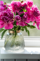 A bouquet of bright pink peonies in a glass vase on the windowsill. Flowers and buds in the vase. Light pink, floral background.