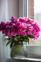 A bouquet of bright pink peonies in a glass vase on the windowsill. Flowers and buds in the vase. Light pink, floral background.