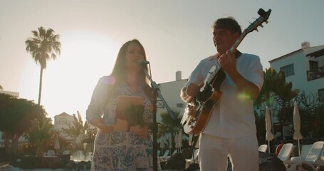 Woman sings and man plays guitar at sunset near resort poolside stage