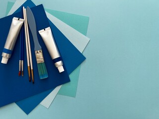 Set of painting supplies including paint tubes, brushes ,and sheets of paper in blue and turquoise...