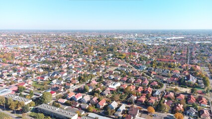 Drone aerial view of a suburban residential neighborhood with rows of houses, gardens, and tree-lined streets under clear blue skies. Peaceful community layout with family homes and green areas.