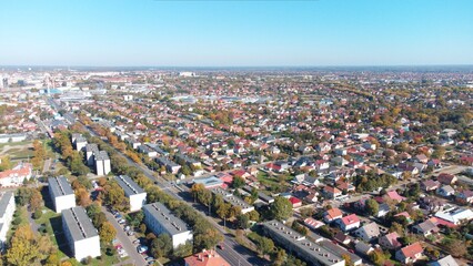 Drone aerial view of a suburban residential neighborhood with rows of houses, gardens, and tree-lined streets under clear blue skies. Peaceful community layout with family homes and green areas.