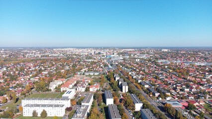 Drone aerial view of a suburban residential neighborhood with rows of houses, gardens, and tree-lined streets under clear blue skies. Peaceful community layout with family homes and green areas.
