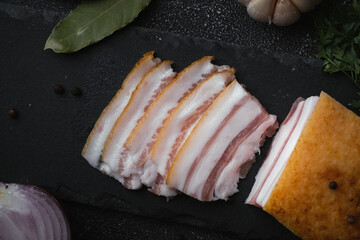 Salted lard bacon sliced on the black concrete background decorated with bay leaf,  garlic and onion. Pork fat meat as healthy food. Top view. Traditional Ukrainian food