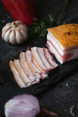 Salted lard bacon sliced on the black concrete background decorated with bay leaf,  garlic and onion. Pork fat meat as healthy food. Traditional Ukrainian food