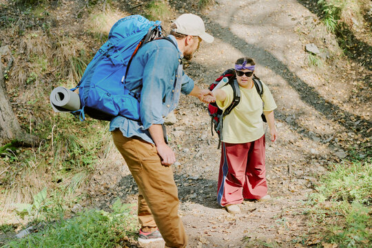Caucasian young adult man helping Caucasian young girl with Down syndrome climb rocky forest trail, both carrying large backpacks and hiking gear, engaging in teamwork during outdoor adventure