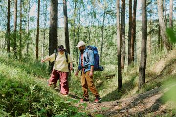 Young Caucasian girl with Down syndrome and young adult Caucasian man hiking together through forest trail, carrying backpacks and outdoor gear, navigating uneven terrain surrounded by tall trees