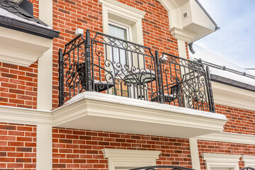 Elegant brick townhouse exterior in winter, a decorative wrought iron balcony with scrollwork, white trim accents, and a large window.  Refined living. Fresh snow on metal chairs and table 