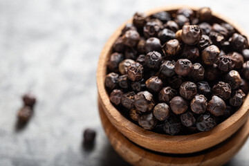 Whole black peppercorns in wooden bowl on light grey surface, sharp focus with shallow depth of field. Concept of spicy flavor and gourmet cooking