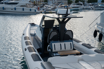 Inflatable boat (RIB) with white and black cushions, featuring a sleek console and dual seats under a T-top with electronics, moored in a marina with other boats in the background.