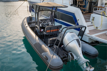 Inflatable boat (RIB) moored next to a yacht, featuring a black hull with cushioned bench seats, a central steering console with gauges and controls, and a large outboard motor at the rear.