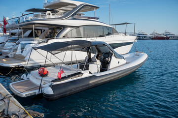 Inflatable boat (RIB) with a black hull and white deck, equipped with a black canvas canopy, a central console, and a UK red ensign flag. It is moored in a marina beside a large white yacht under clea