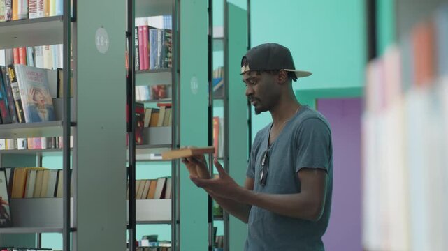 Young man in casual t shirt and cap reaches for book on library shelf, preparing to open it, standing in bright teal walled corridor lined with books, focused and engaged in academic environment
