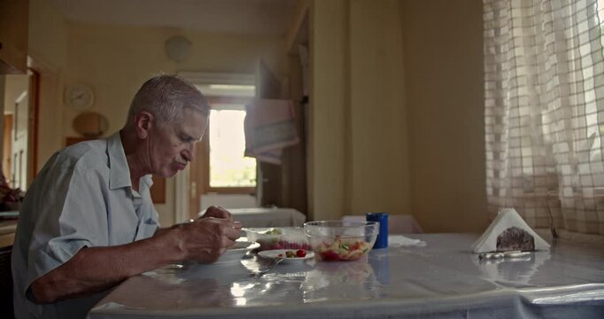 An elderly man sits at his kitchen table, enjoying a healthy meal of salad and soup. He appears focused and content, highlighting themes of independent living and a calm, domestic routine at home.