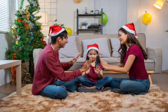 Asian parents and young daughter wearing Santa hats, playing clapping games on the floor, celebrating Christmas at home