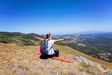 Naklejka premium Female hiker with backpack sitting on mountain peak enjoying panoramic landscape view of rolling hills and valleys.