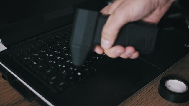 Angled shot of a hand operating a portable electric air blower to remove dust from black laptop keyboard keys. Modern cleaning gadget for device maintenance on a wooden desk or workshop setup.