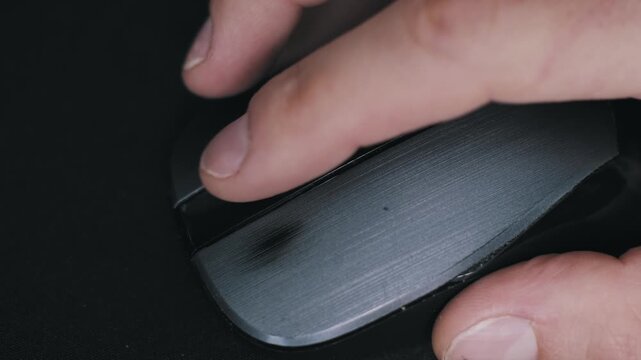 Top down macro shot of a male hand operating a black computer mouse. A finger scrolls the metal wheel and clicks the left button on a dark mousepad surface, browsing the internet for online work.