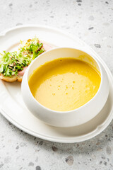 A bowl of creamy orange soup, likely pumpkin or squash, served on a white plate with an open-faced sandwich. The meal is presented on a textured grey background, shot from an elevated angle with natur