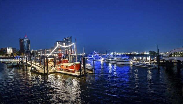View of illuminated ships on the Elbe promenade with restaurant Das Feuerschiff LV 13, City Sporthafen, behind Speicherstadt with Elbphilharmonie and Columbus Haus, blue hour, Hamburg, Germany