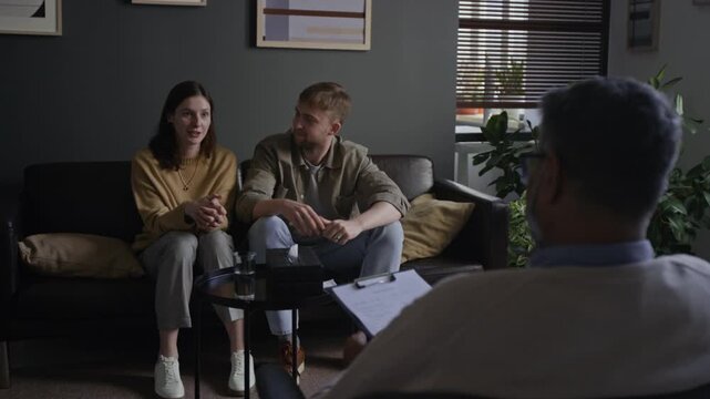 Over shoulder shot of smiling young couple embracing sitting on couch and listening during family therapy session with mature Indian man as therapist in foreground, copy space - Powered by Adobe
