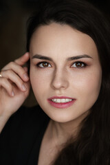 Fototapeta premium oman with dark, wavy hair and freckles, wearing a black top, touching her forehead while looking to the side in a dark indoor portrait with soft lighting