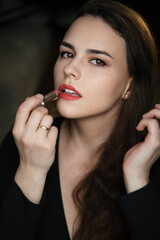 
Woman with long dark hair and freckles, applying red lipstick while looking at the camera, wearing a black top in a darkly lit indoor portrait setting